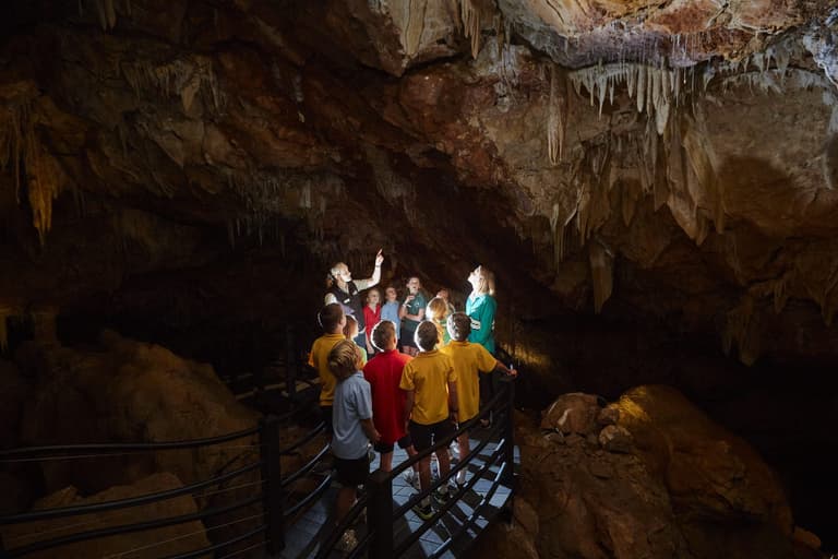 Group of children in uniform yellow, red, green and blue shirts and a tour guide inside a gave looking up to where torch is shining.