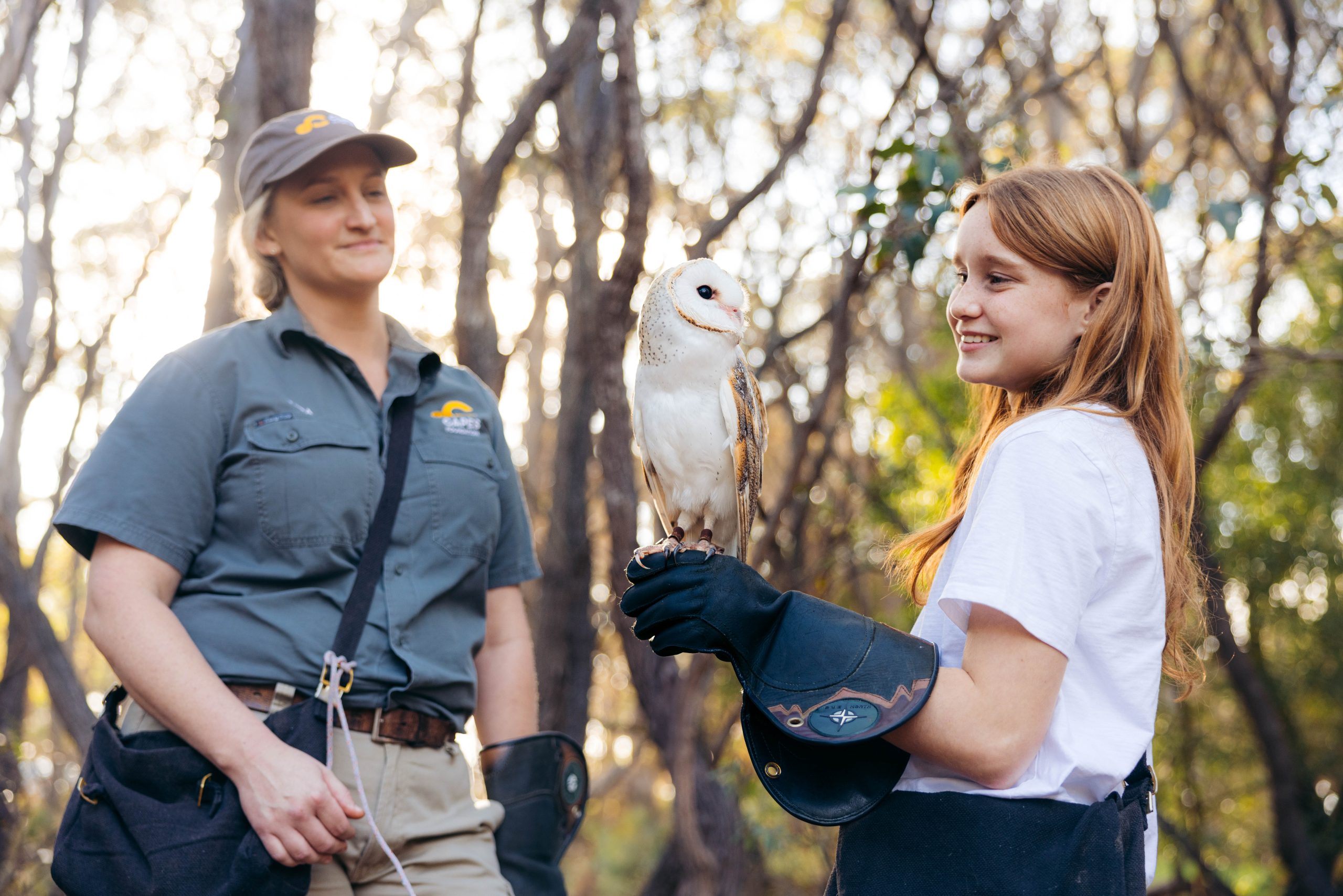 A smiling girl, with a Barn Owl perched on her glove, with a Capes Raptor Centre staff member be her side