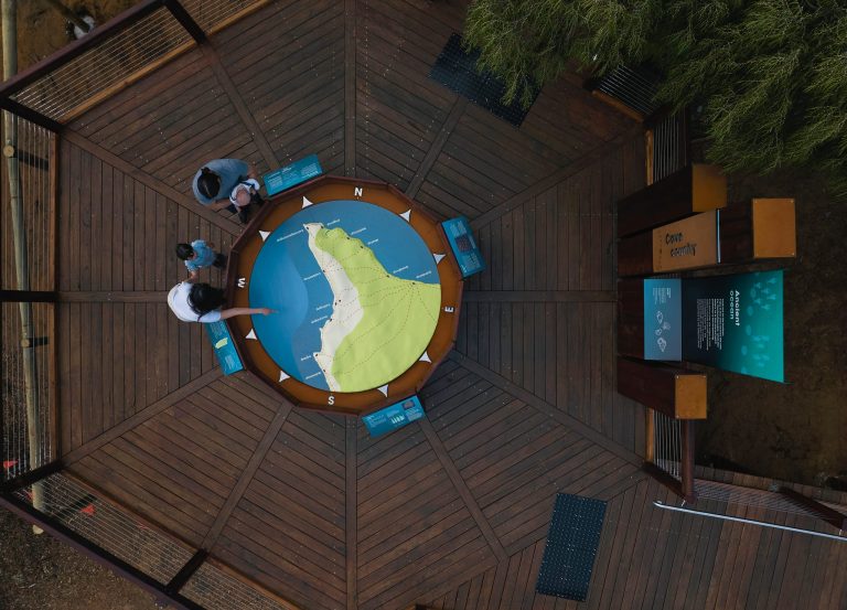 Aerial view of a family standing on a timber viewing deck at Ngilgi Cave’s Ancient Lands Experience, gathered around a large circular map of Cape Naturaliste with interpretive panels and bushland surrounding the platform.