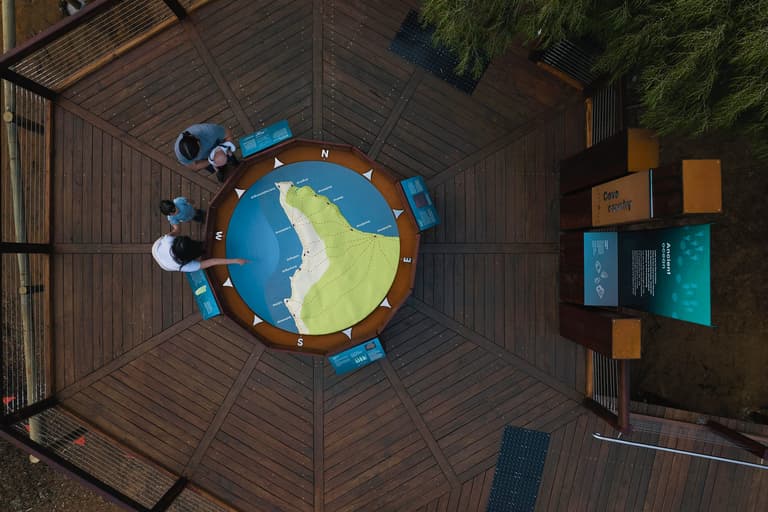 Aerial view of a family standing on a timber viewing deck at Ngilgi Cave’s Ancient Lands Experience, gathered around a large circular map of Cape Naturaliste with interpretive panels and bushland surrounding the platform.
